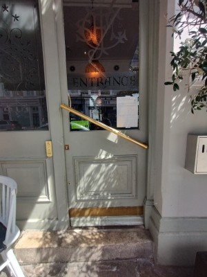 One step leading up to single door. Wooden door with handle diagonally across door. Push open from the left.