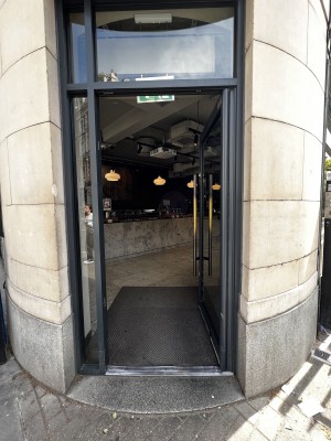 From pavement, facing straight. Glass door with black frame. Long vertical handle on the left. Push to open.