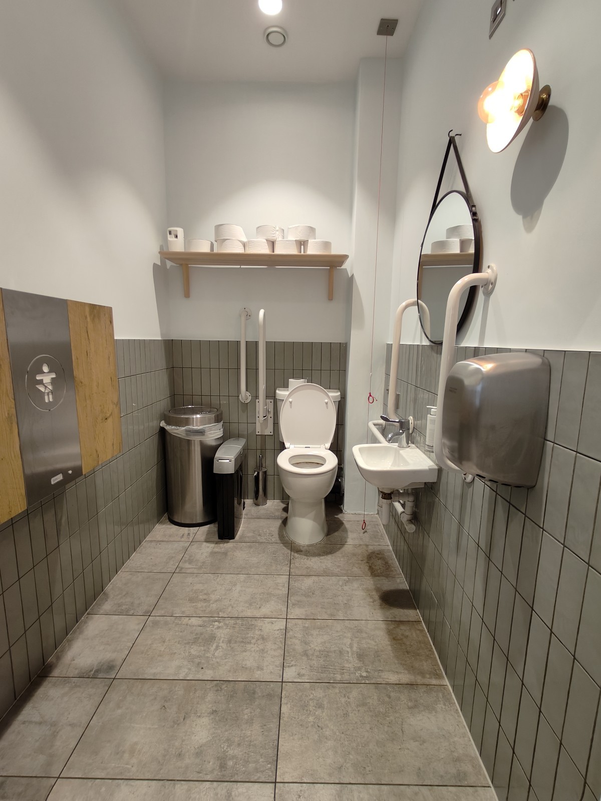 From door facing straight. Baby change table on left. Toilet at back. Pull cord to its right. Sink on right wall.