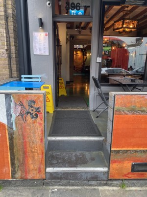 From sidewalk facing venue entrance. Barriers on left and right. Steps ahead leading to door at back.