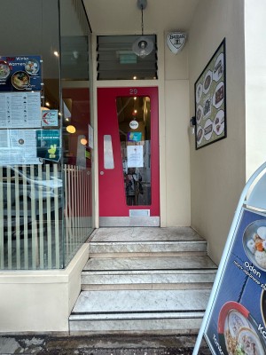 Steps up to red wooden door with glass panel at back. Push from left to open. A-frame sign on right.