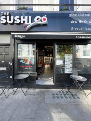 From pavement, facing straight. Path leads to black metal-framed glass door ahead. Push black handle on left side to open. Seating to left and right.