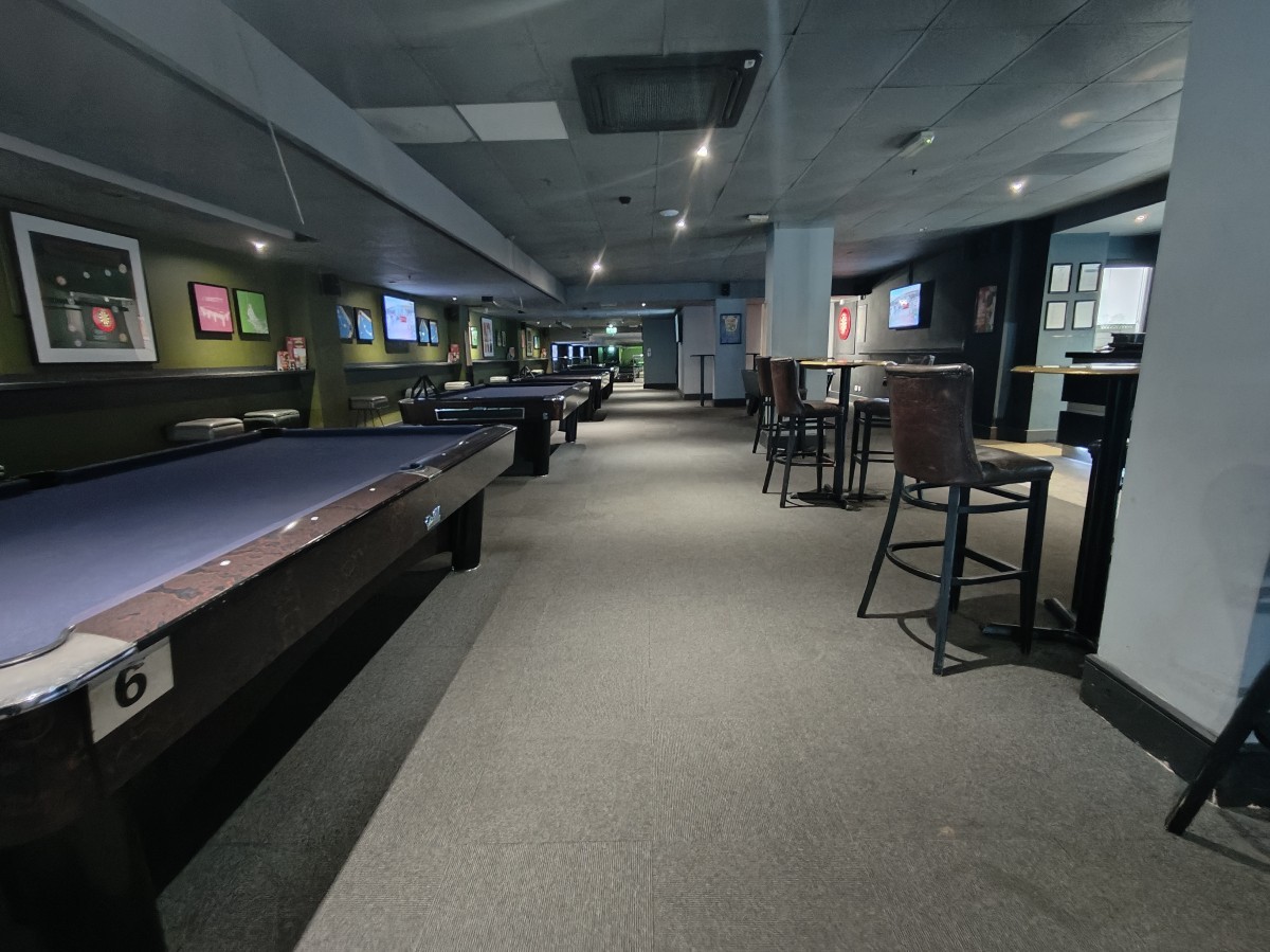 From before pool tables facing right. Pool tables along left. Path ahead to back area. Stools continue along right.