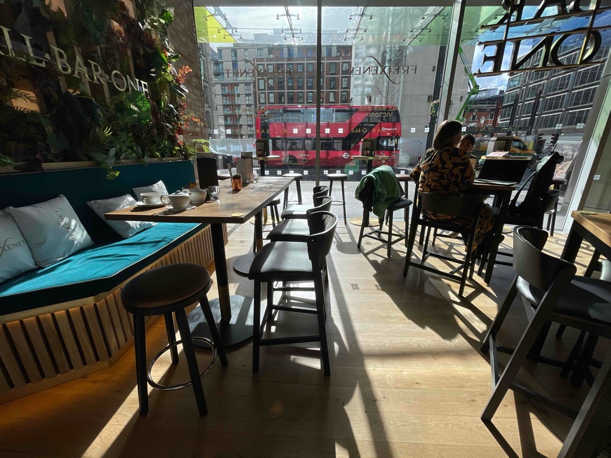 From end of bar counter, facing right. High half booth along left. Path ahead, going straight. High tables spread out at right. Glass wall at back.