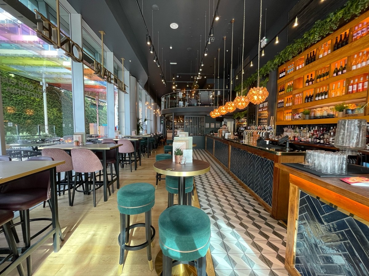 From left of entrance, facing left. Bar counter along right. High table ahead. Paths on left and right. High tables along left. Glass wall far left.
