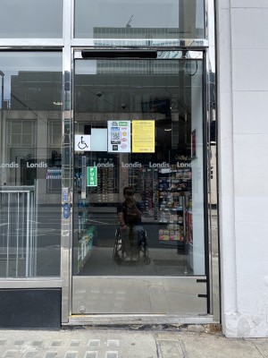 Main entrance with single step and glass, manual door. Door opens inwards with push handle on the left. Disabled assistance doorbell on right.