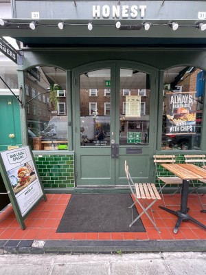 From sidewalk facing venue. Entrance straight ahead. Elevated area in front. Seating to immediate right of door. Sandwich board on left.
