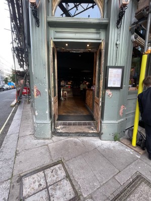 One concrete step before entrance. Walls on left and right. Wood-panelled glass doors after step. Pull brass handles at centre to open.