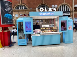 Retail area/entrance of Lola’s cupcakes. Retail counter below, up top double blue doors open out with screens on.