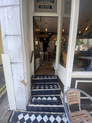 From pavement, facing straight. Steps ahead. Handrail to left. Glass door with brown frame after stairs. Gold handle to right. Push to open.