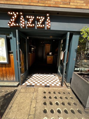 From pavement, facing straight. Glass doors with green frame ahead. Verticle metal handles in centre. Pull to open.