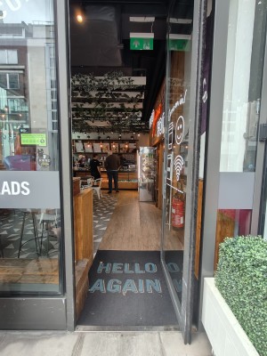 Facing grey framed single glass entrance door. Push silver metal panel on left of door to open.