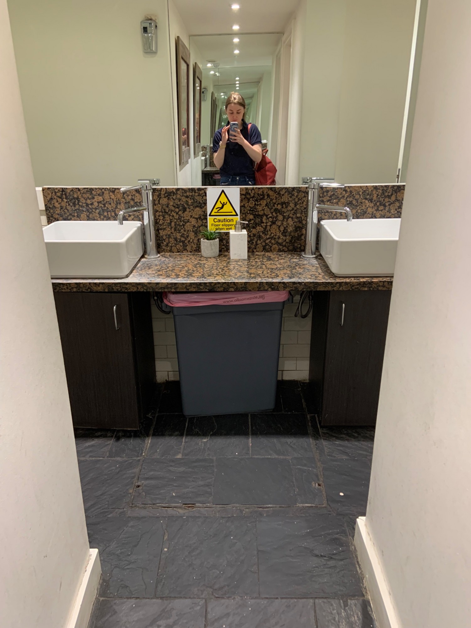 View from doorway to bathrooms looking right. Pathway in centre. Counter with sinks along back wall with open bin underneath. Pathway in front of sinks leading to bathrooms to left and right.