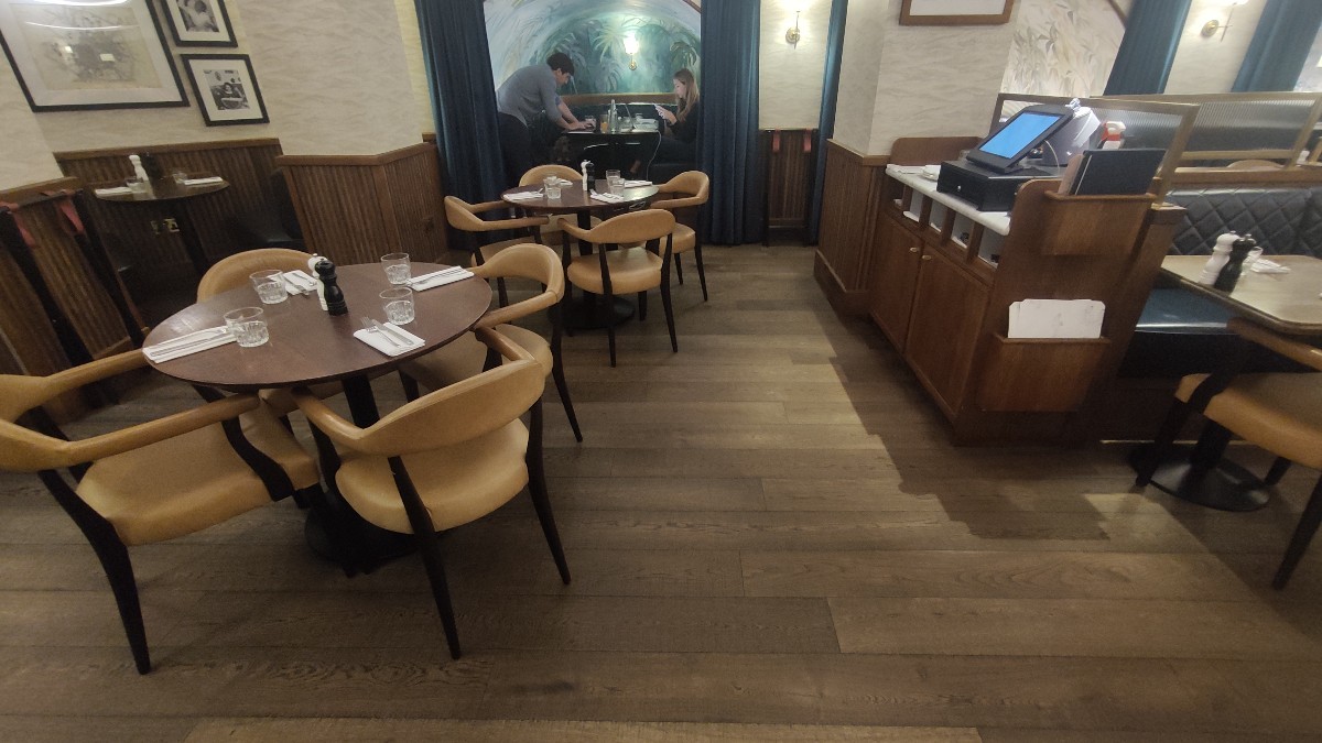From bottom of staircase facing left. Tables and chairs along left. Path on right. Booths with curtains at back. Staff counter at back right.