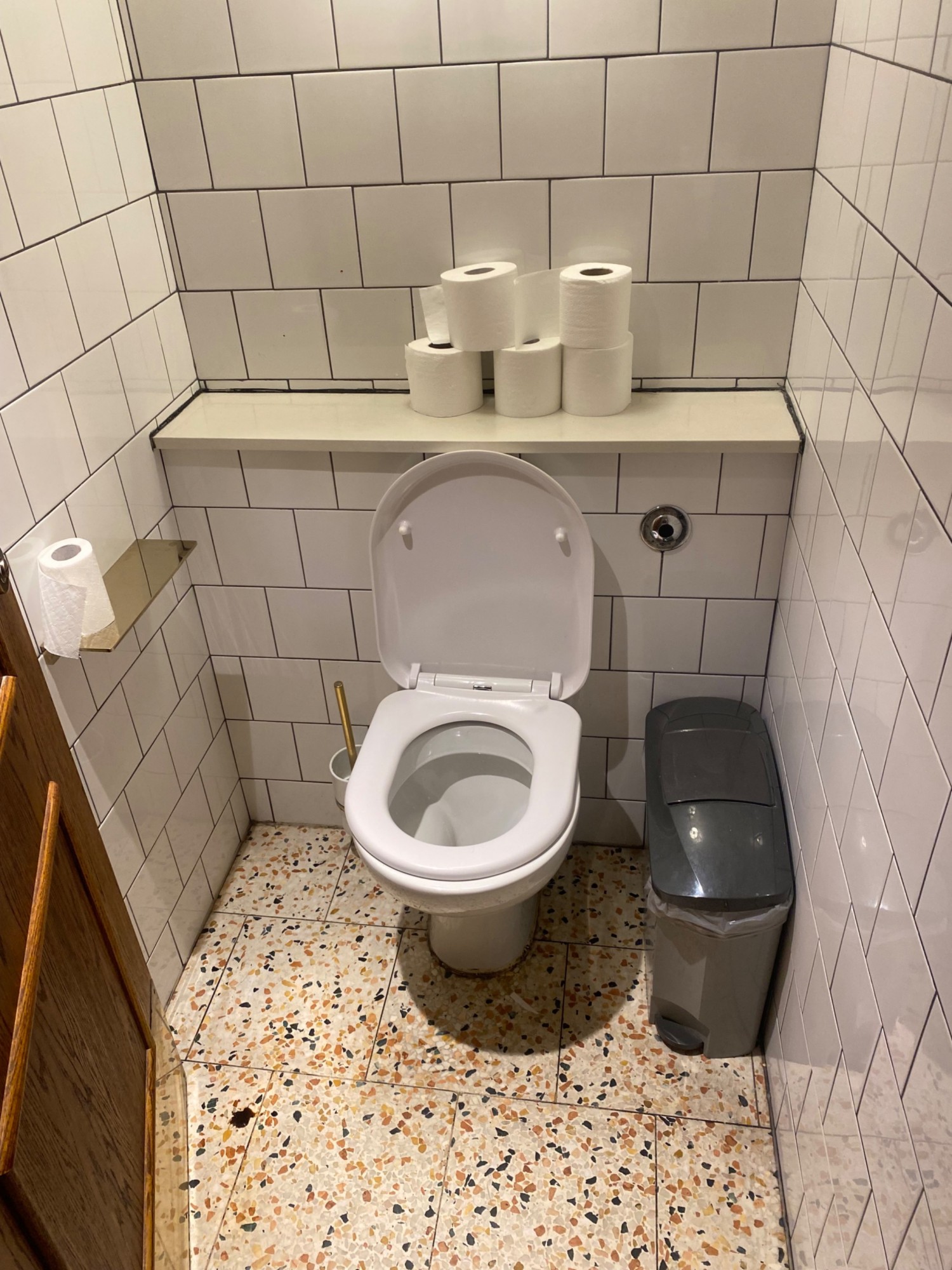 From door facing straight. Toilet ahead. Sanitary bin on right of it. Standing height shelf at back.