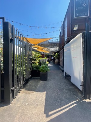 Large black iron gate ahead. Push from right to open. Plant pots ahead on left and bench on right after gate.