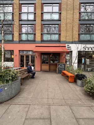From sidewalk facing venue. Opening ahead between planters. Path to entrance on back with benches along both sides.