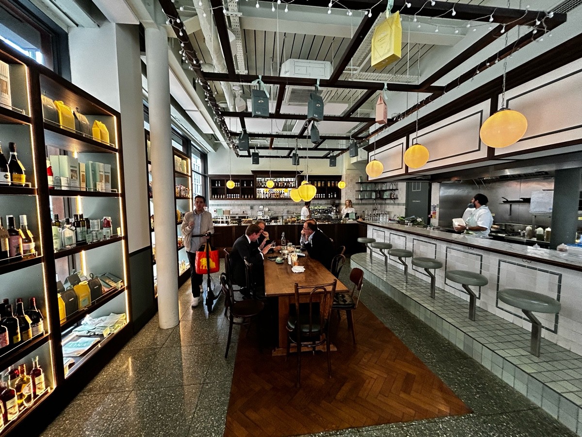 From entrance, facing left. Open shelves along left. White pillar back left. Table with chairs ahead. Narrower paths either side. Bar along right.