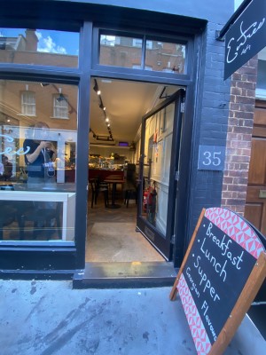 Single glass door with black wooden frame. Push brass handle on left to open. Doormat after entrance. A-frame right.