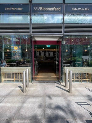 Double glass doors in glass frame. Push silver metal vertical handles at centre to open. Metal poles on left and right of path before entrance.
