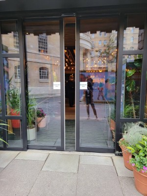 Main entrance looking straight. Flower pots left side. Main door center. Two glass automatic doors. Flower pots left.