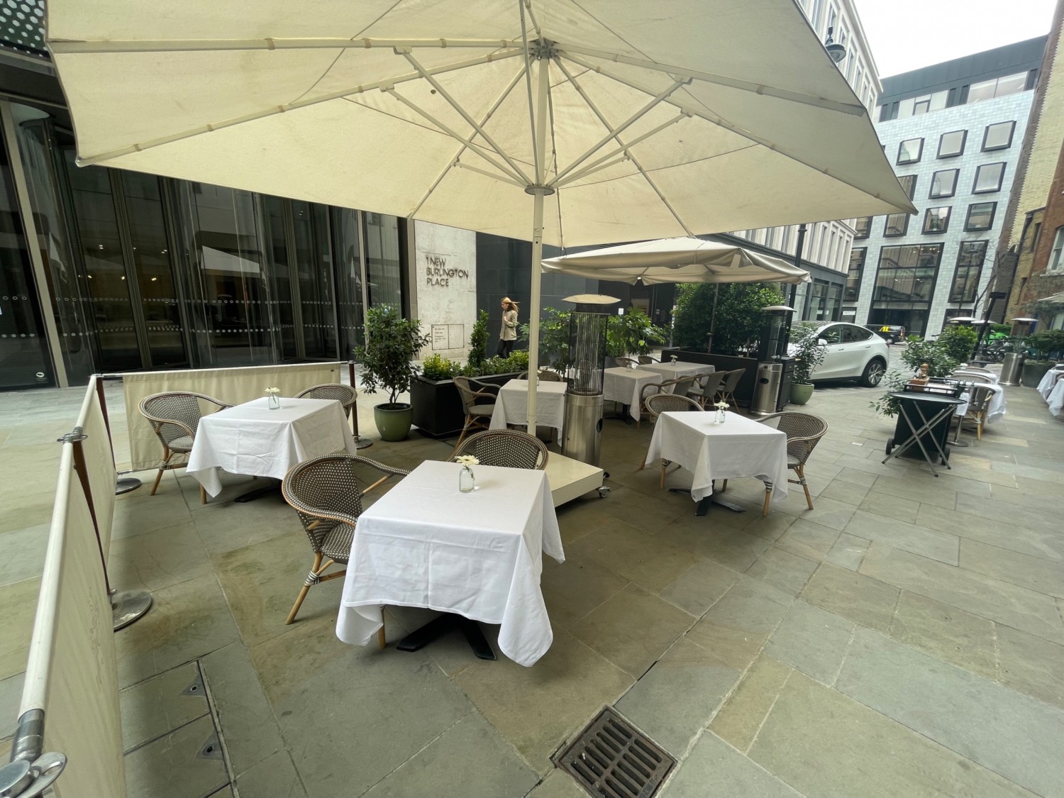 Looking to the left, from entrance to outdoor area. Tables and chairs in centre and to the left. Large umbrella base obstructing path in centre. Pathway leading to further seating to the right.