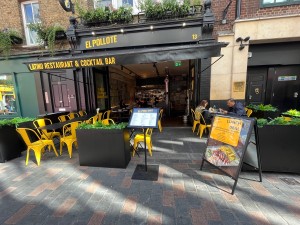 From pavement facing venue. Path ahead to main entrance proceeds through outdoor seating. Sandwich board right. Menu sign left.