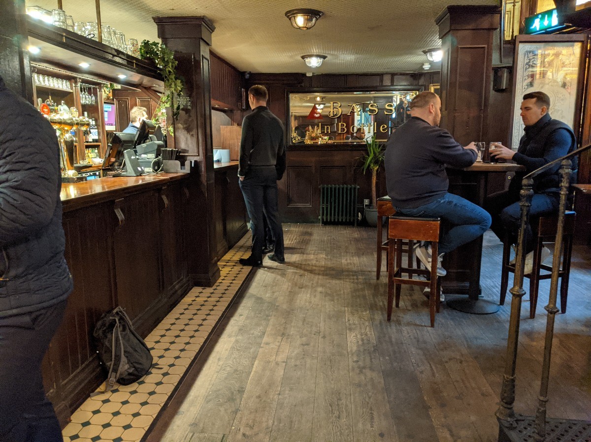 From pillar facing right. Bar along left, path to alternative entrance ahead. Stairs to upstairs on right. High tables with stools behind.