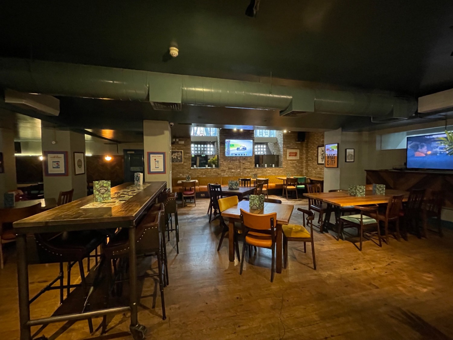 From middle of downstairs area looking straight. High table on left. Pathway to right. Tables and chairs on right. Half booth seating along back wall.