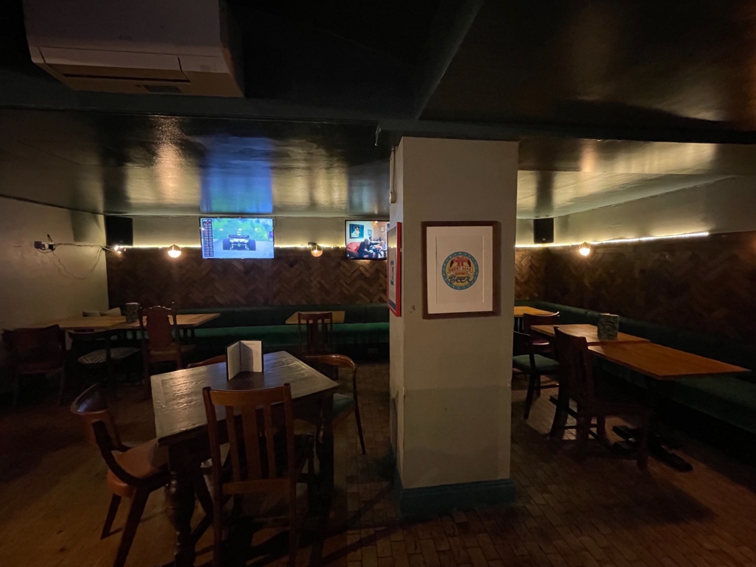 back of downstairs area looking left. To Centre left pillar. Half booth along right wall with tables and chairs. Half booth seating along back wall.