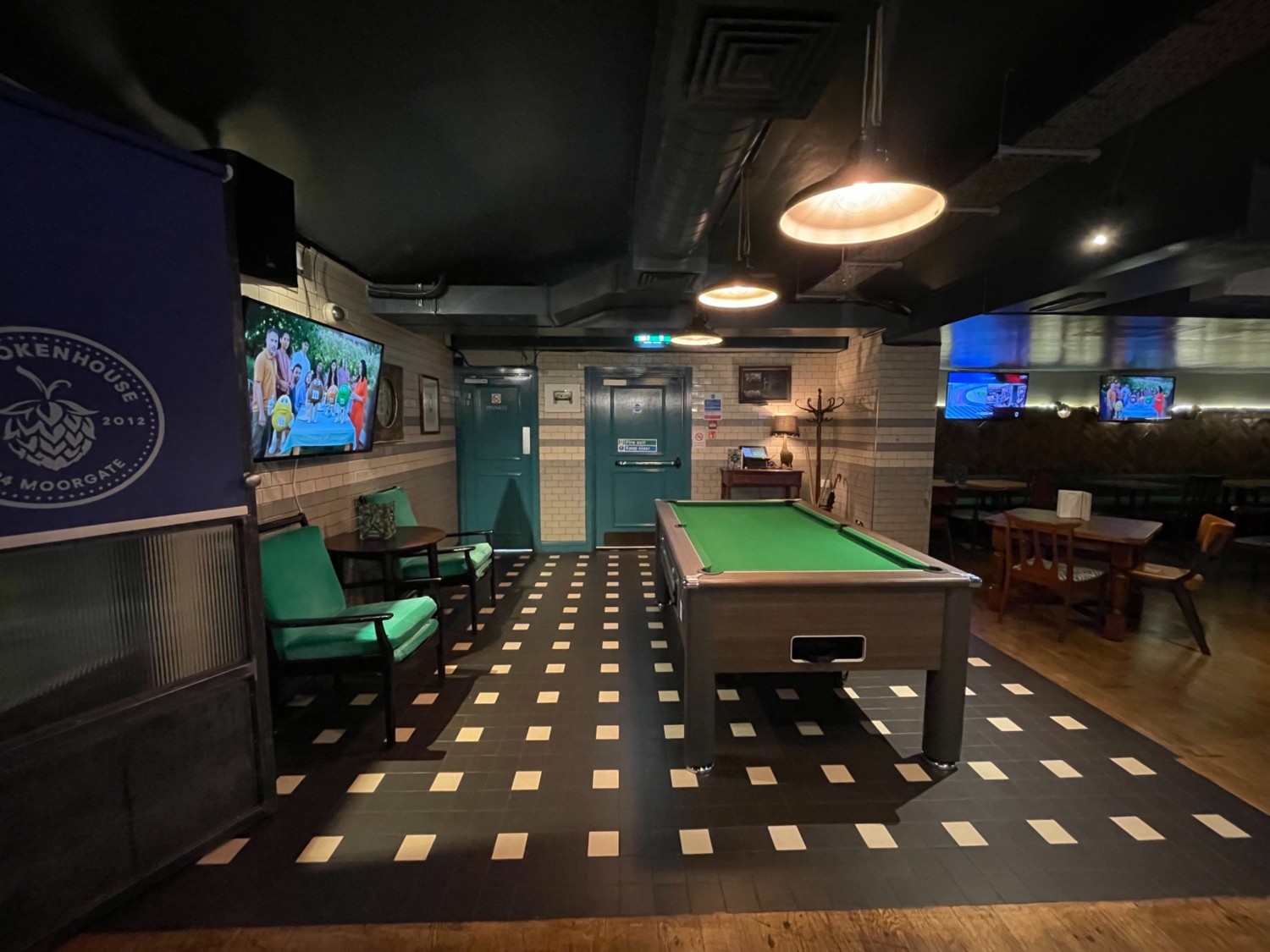 From middle of downstairs area looking left. On left wall two arm chairs. Pathway. Pool table to the right. Tables and chairs in the right corner.