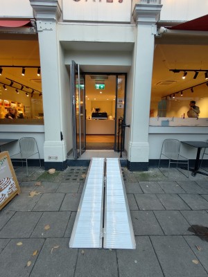 Facing entrance. Ramp on request ahead to black framed glass door. Pull silver metal handle on right to open. Black handrail on right after ramp.