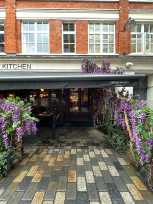 From sidewalk facing venue. Path to entrance ahead. Planters along right and ahead on left. Seating in back left.