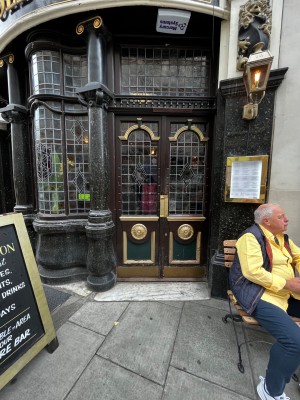 Double glass doors with brown wooden frames. Push gold metal panels from centre to open. Sandwich board left. Outdoor seating right.