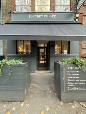 From sidewalk facing venue. Path to entrance ahead. Large planters either side of path.