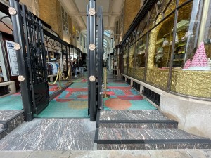 From sidewalk facing arcade leading to venue. Ramp ahead on left. Stairs ahead on right. Entrance in back right.
