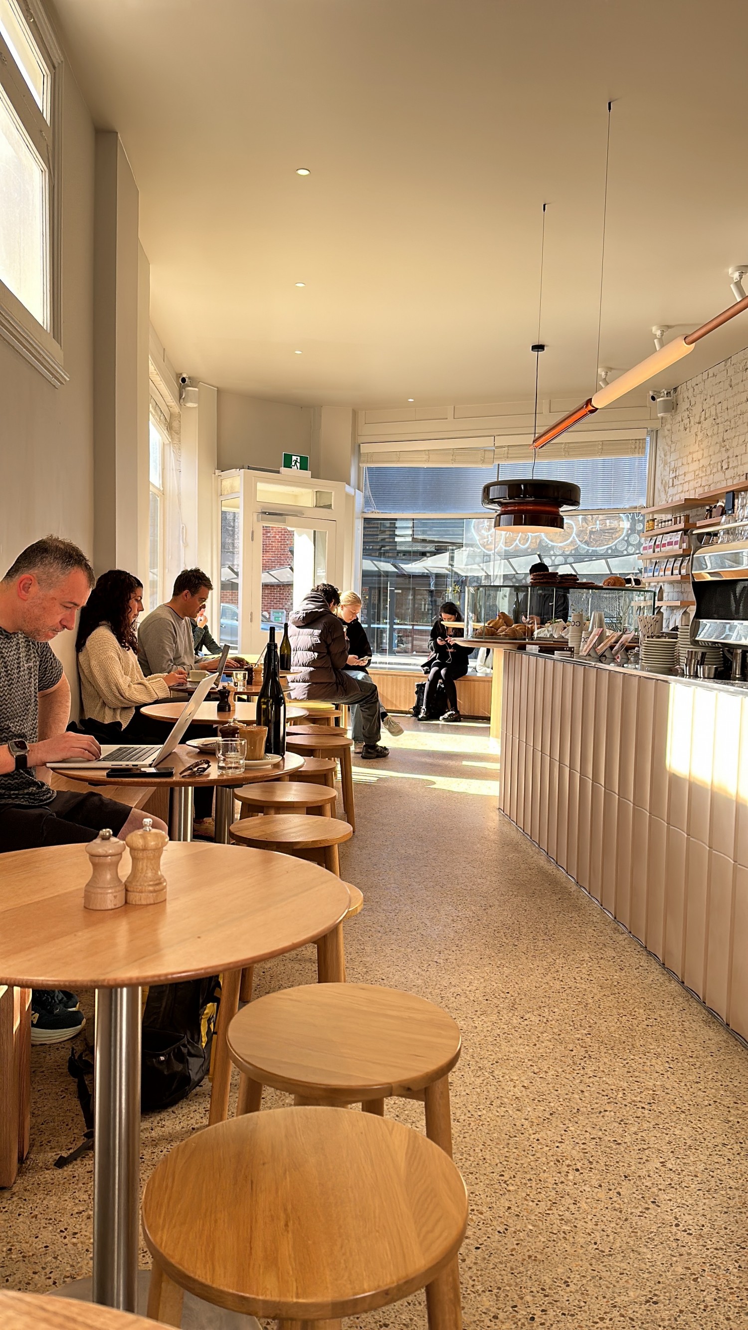 Image of indoor of cafe taken from rear of cafe. Stools and tables running down left side and front door on far left. Counter running down right hand side. Front in background set in front of window.