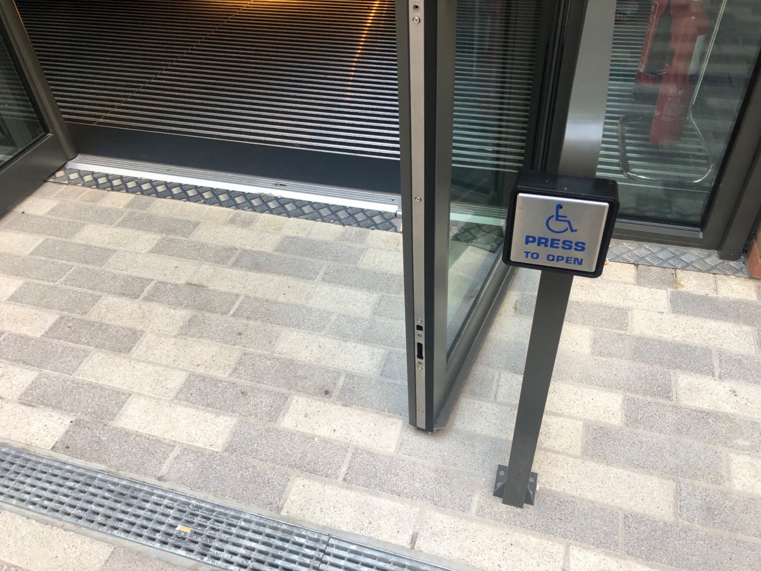 Seated height push button mounted  on pillar at front entrance doors. Floor outside is smooth bricks and there is a small metal bump at entrance way.