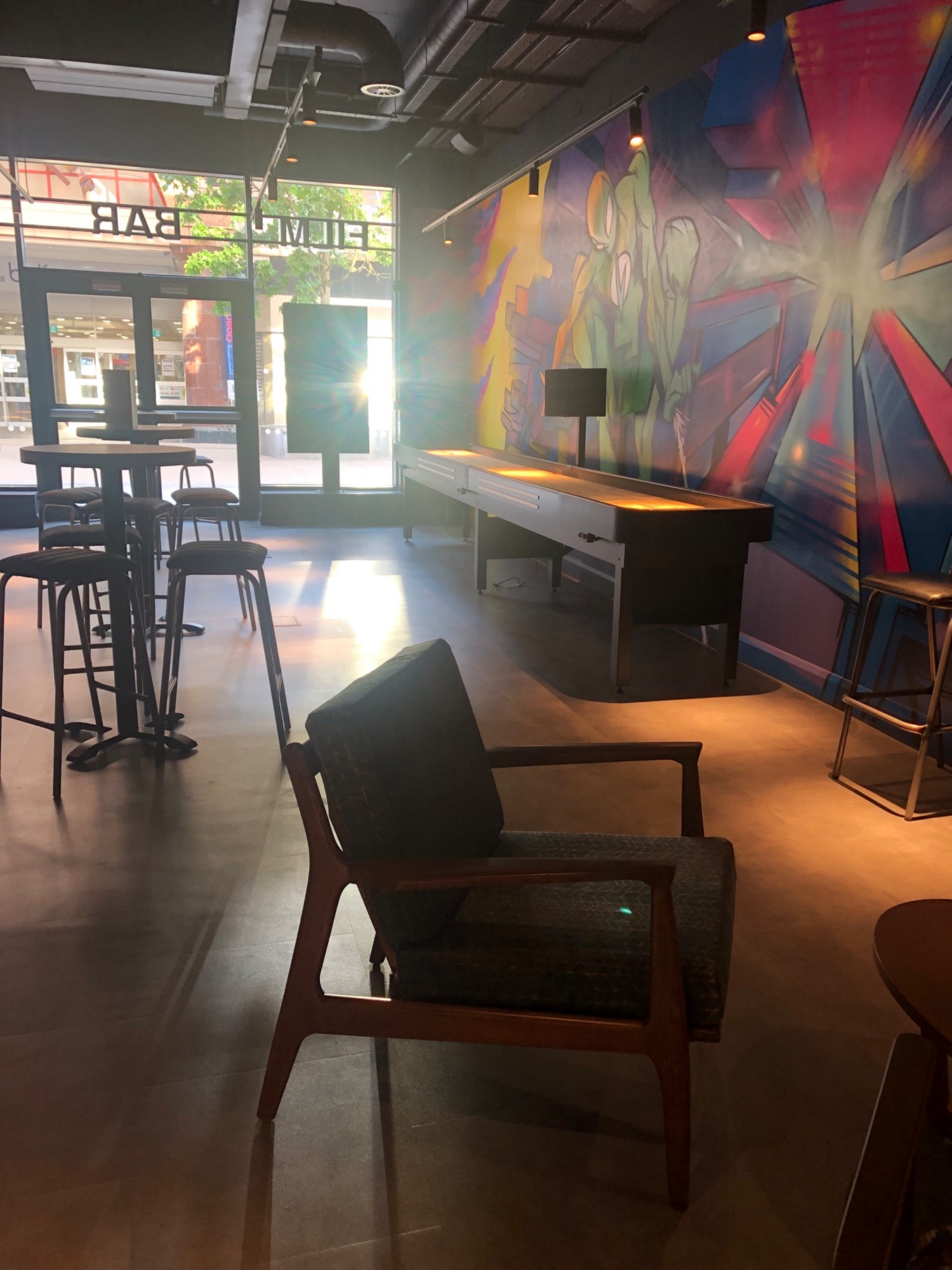 Mixed seating of high bar stools at high table and low lounge chairs. Shuffle board by dark wall. Wide walkways between chairs. Glass windows at back