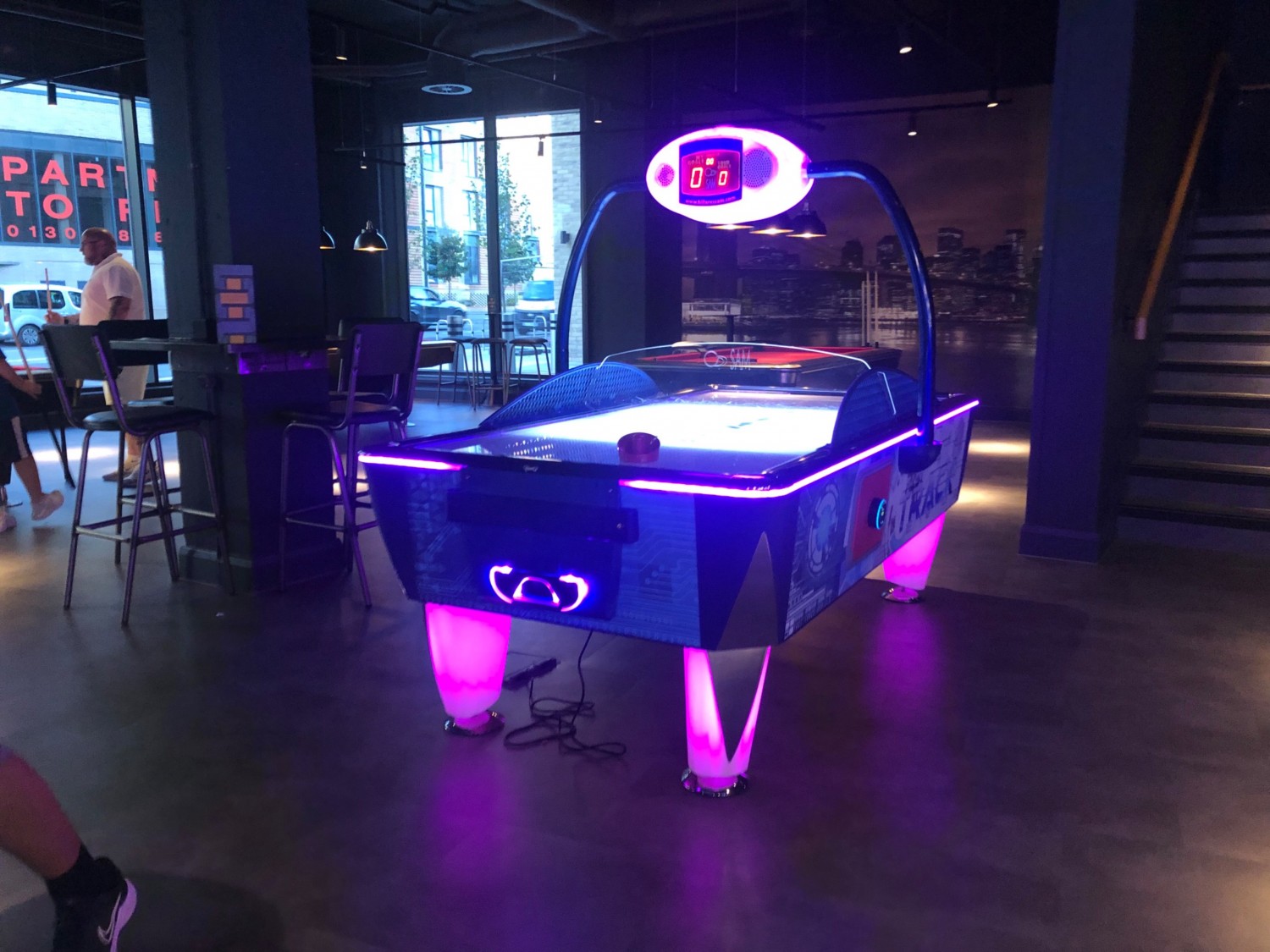 Neon glowing air hockey table in arcade area, one trailing electrical wire underneath and high stools and table nearby.