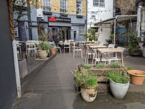 From pavement facing venue. Ramp ahead leading to outdoor seating area. Planters on right, one step up right of planters.
