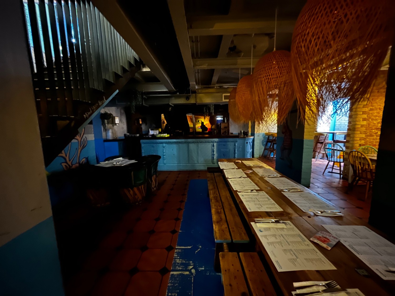 Continuation of downstairs area back past long table with benches. Bar counter at back of space, table with chairs on left. Area continues to left.