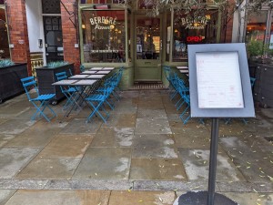 From pavement facing venue. Menu stand on right, outside seating on left and right behind. Door to venue at back.