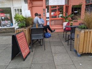 From pavement facing venue. A-frame sign on left, planter behind. Planter on right. Door to venue back right.