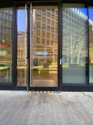 Glass doors with black frame ahead. Push from handle in centre to open. Assistance doorbell to right of doorway.