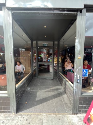 From pavement facing venue. Open doorway ahead. Assistant button on right. Path behind.