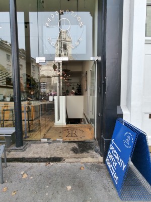 Facing double glass doors. Push silver vertical handles in centre to open. Doorbell for assistance on right.