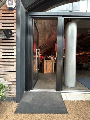 Single glass door with black metal frame and silver metal vertical handle along right. Push to open. Silver pillar to right of door.