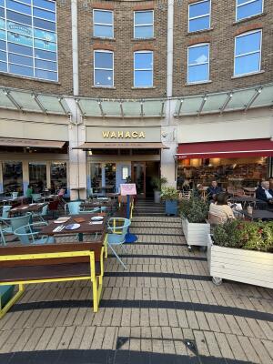 From sidewalk facing venue. Planters along right. Path down centre to entrance at back. Outdoor seating to left.