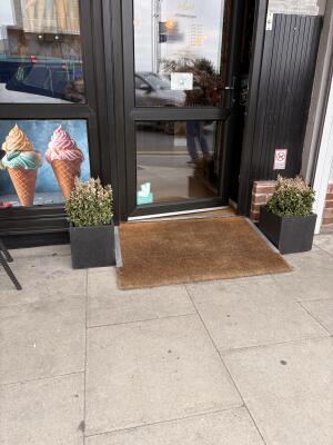 Black metal-framed glass door. Pull from handle on right to open. Brown doormat and planters on each side of entrance.